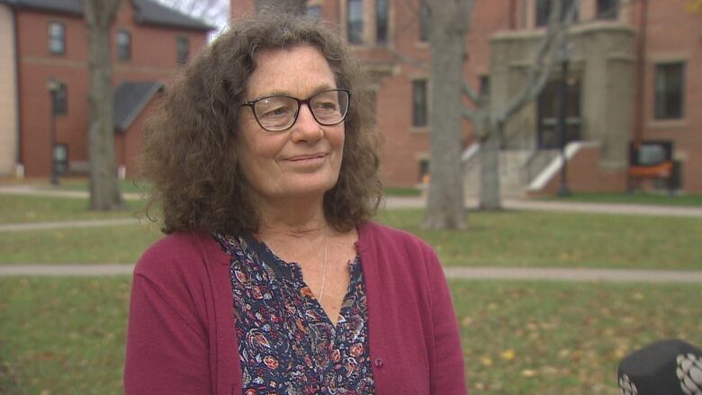 A woman with long brown hair and glasses stands on a university campus.