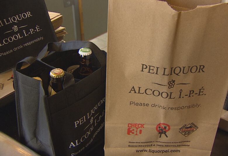 Paper bags with P.E.I. Liquor Control Commission branding sits on the conveyor belt of a check out counter. A cloth bag sits beside it with glass bottles inside. 