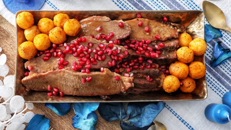 Overhead shot of a rectangular serving dish with brisket and mini potatoes with pomegranate arils on top. The decor on the table is blue and white. 