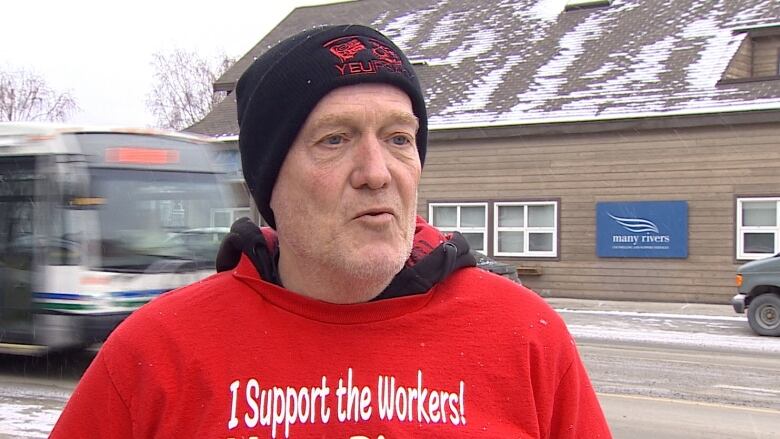 A man in a toque and red shirt stands on the side of a street.