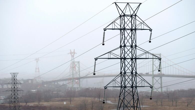 Power line towers stand in a field, with a bridge in the background, on a foggy day.