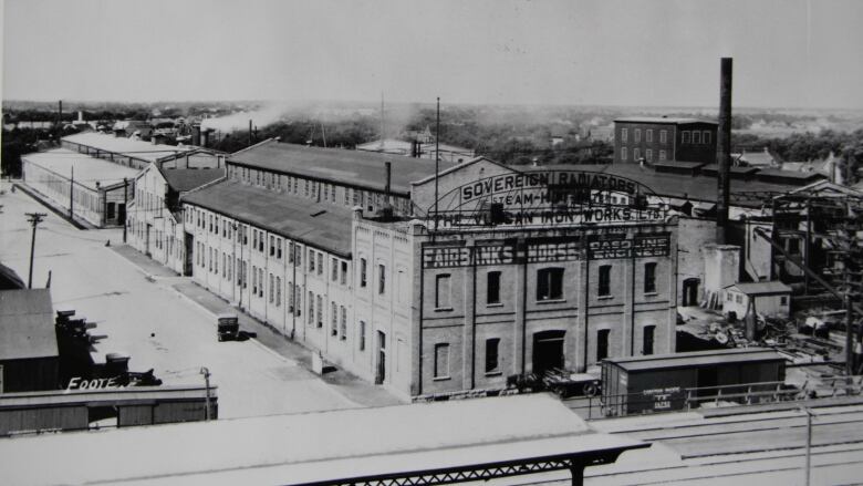 A black-and-white photo shows a complex of buildings next to a railway line.