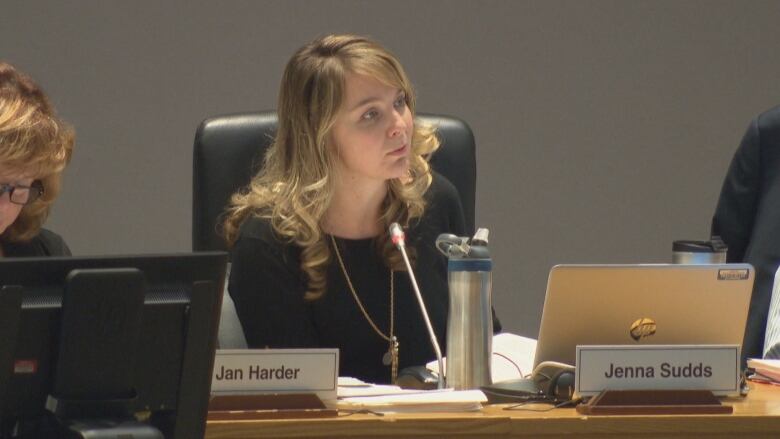 Jenna Sudds sits at a city council table and speaks into a microphone.