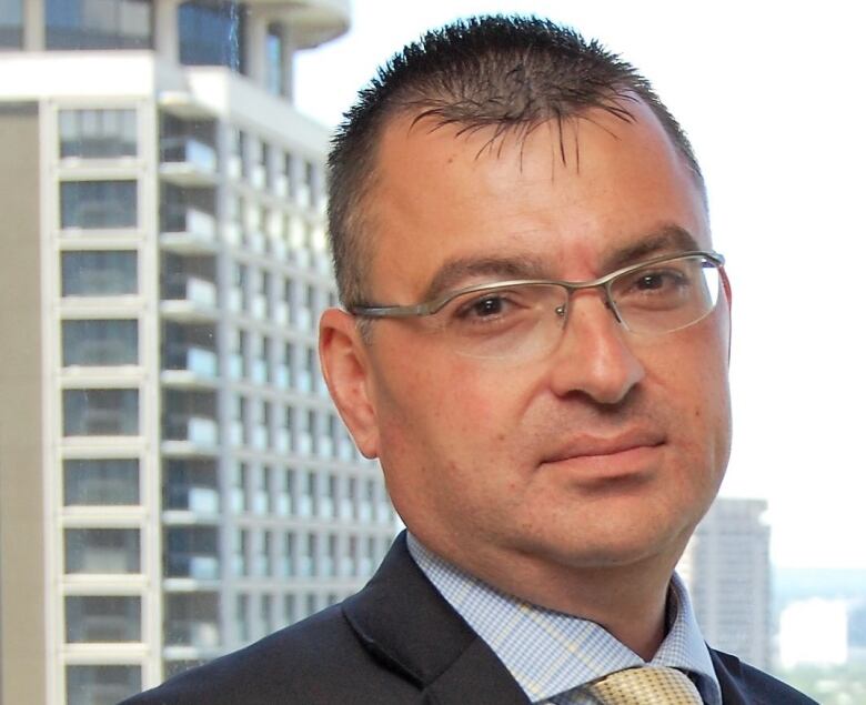 A man with short hair and glasses stands in front of a window with a high-rise building in the background.