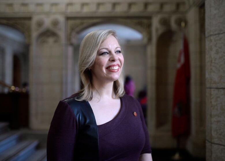 A smiling woman with blond hair looks off camera. She's wearing a purple dress.