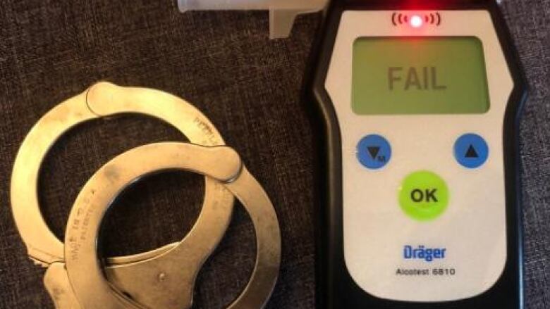 A pair of handcuffs lie on a table next to an alcohol breathalyzer that reads 'fail' on the screen.