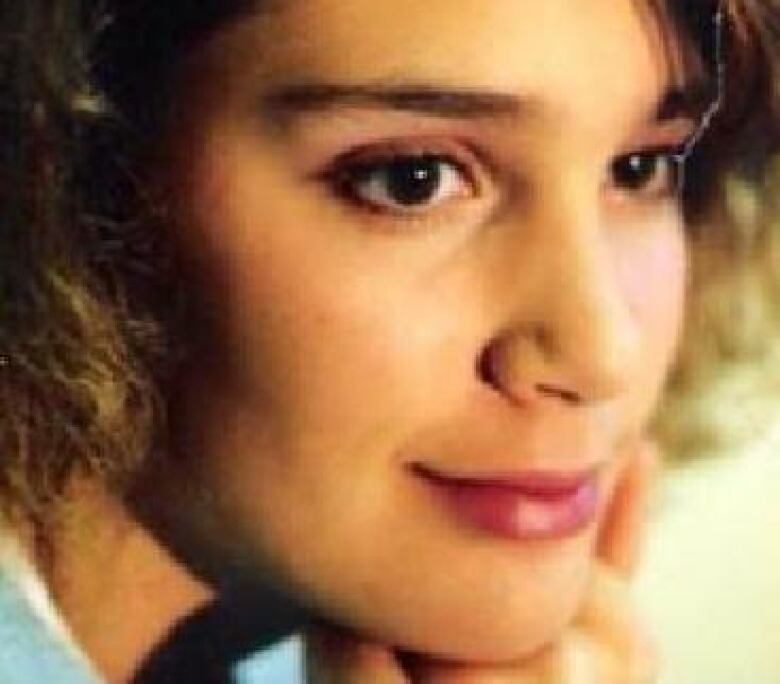 A young woman with curls in her hair and her chin resting on her fingers smiles in a closeup of her face.