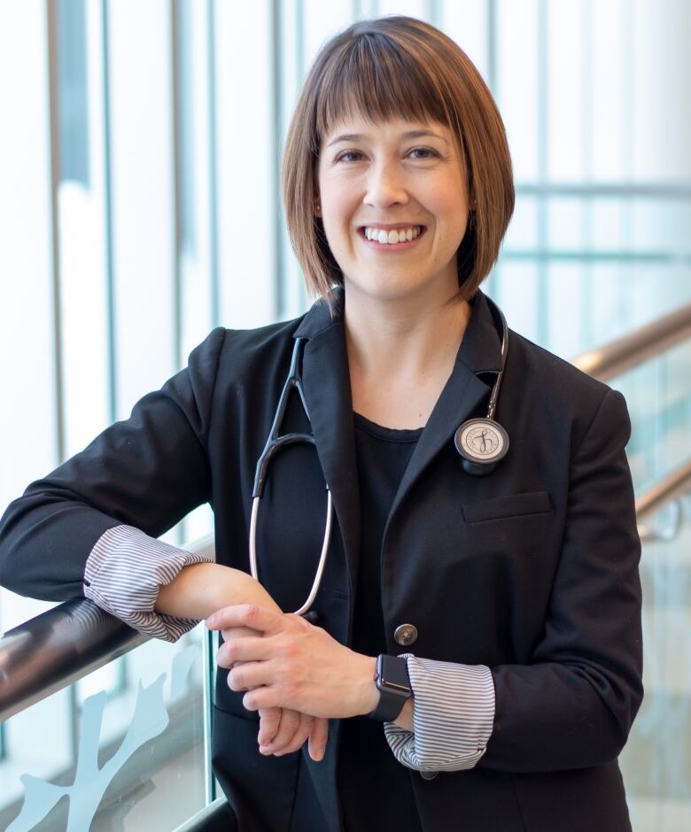 Woman with chin-length dark brown hair wearing black top and suit jacket, with a stethoscope around her neck, stands next to window.