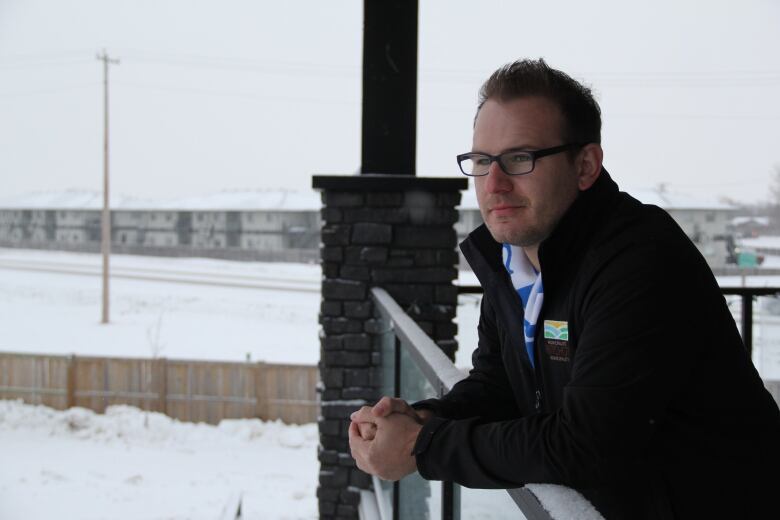 A man with brown hair and black-framed glasses leans on the railing of a balcony, looking out over a snowy field.