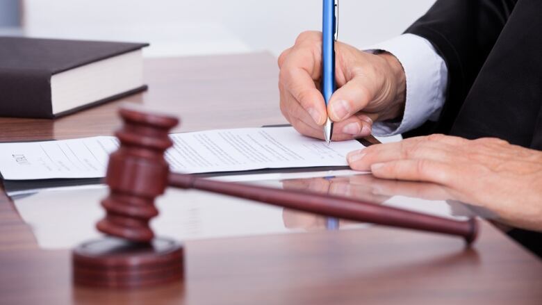 A close-up photo shows a gavel on a desk and the hands of a man writing in a document.