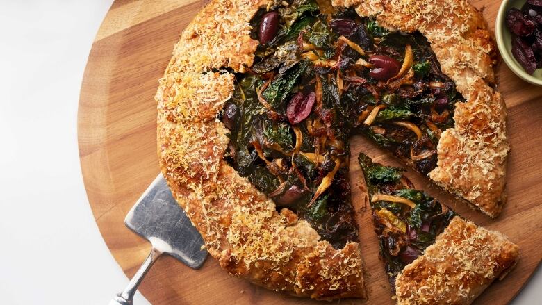 A galette on a wooden cutting board. It's filled with mushrooms, kale and olives. 