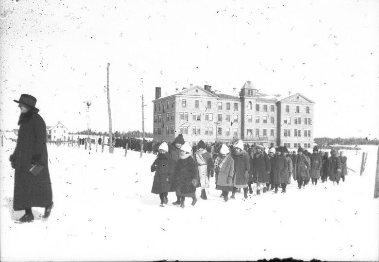 A black and white photo shows children in coats and hats walking through the snow in front of large brick building, following a person wearing a dark coat and wide-brim hat.