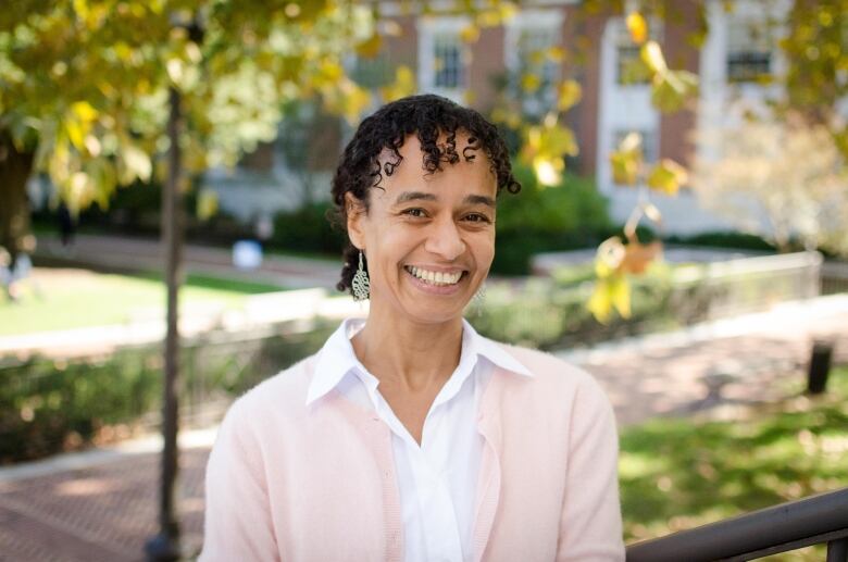 a Black woman wearing a white blouse and pink cardigan smiles at the camera outside a university building