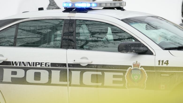 A white police car with black lettering is seen close up and from the side. Blurred yellow police tape is seen in the foreground.