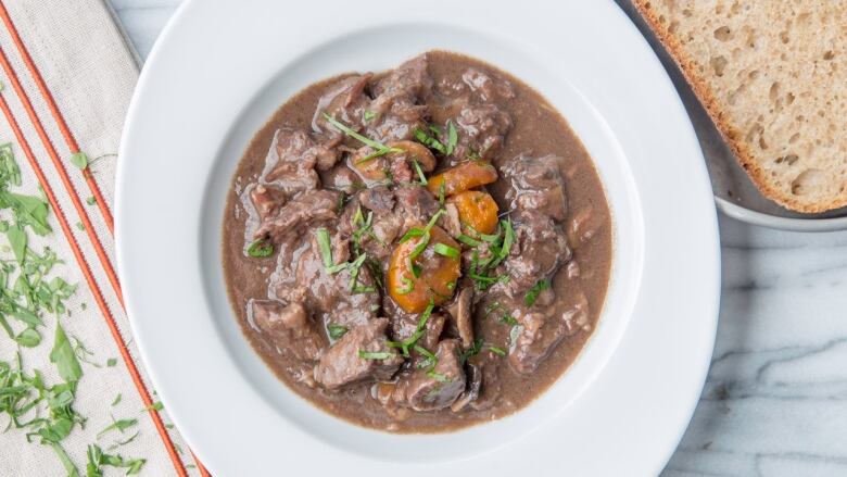 Overhead shot of a bowl of Beef Bourguignon on a marble surface with a slice of bread and a cloth napkin next to it. 