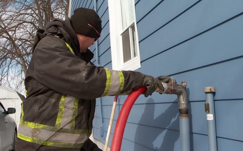 A man in a heavy coat delivers home heating oil from a large hose to an intake pipe on the side of a house in P.E.I. 