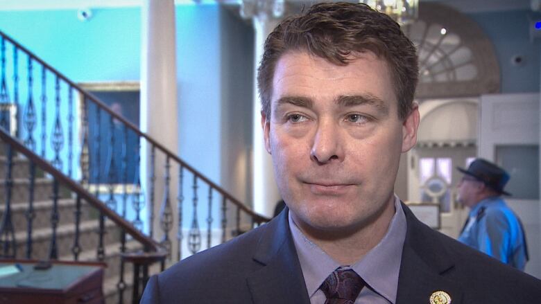 A white man with short brown hair wears a suit and tie in the lobby of Province House, with a staircase behind him