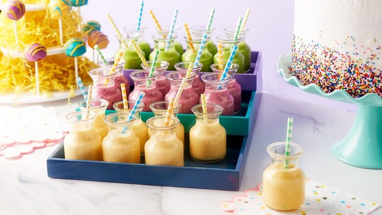 3 colourful trays stacked on a table at a birthday party. the trays contain small glasses with yellow, pink and green smoothies in them. 