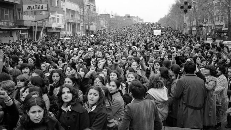 A black and white photo shows a huge crowd of women protesting in Iran.