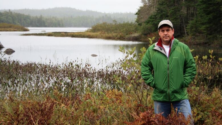 A white man stands in a marshy area in front of a lake wearing a cap, jeans and a green windbreaker