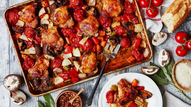 Overhead shot of a sheet pan with roasted chicken thighs, tomatoes, and pieces of mushrooms, red pepper and onion on it. It's sitting on a grey wooden surface with the fresh ingredients scattered around the pan. 