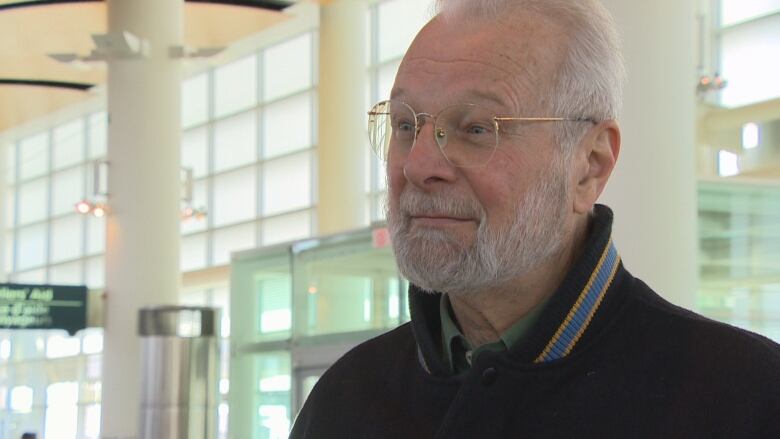 A man with white hair and glasses looks at someone off-camera in an airport.