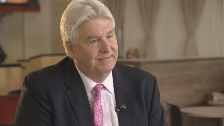 A man with white hair wearing a suit and pink tie sits and smiles into the camera.