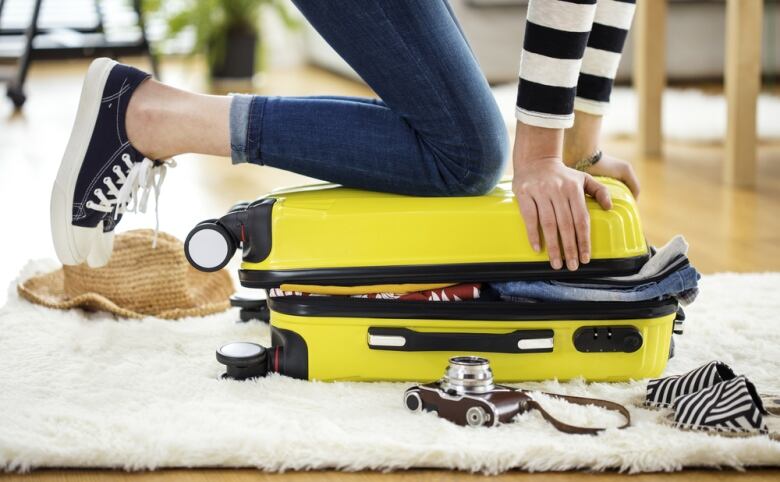 A woman's hands and knees are shown on top of a small, overstuffed yellow suitcase.