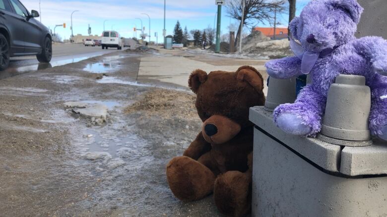 Two teddy bears, one brown and one purple, have been placed at the base of a light pole next to a busy street.