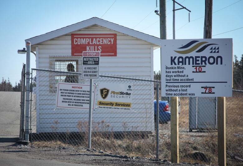 A man is seen inside a guard house behind a chain link fence with a sign saying complacency kills and another saying the mine worked 10 days without a lost time incident.