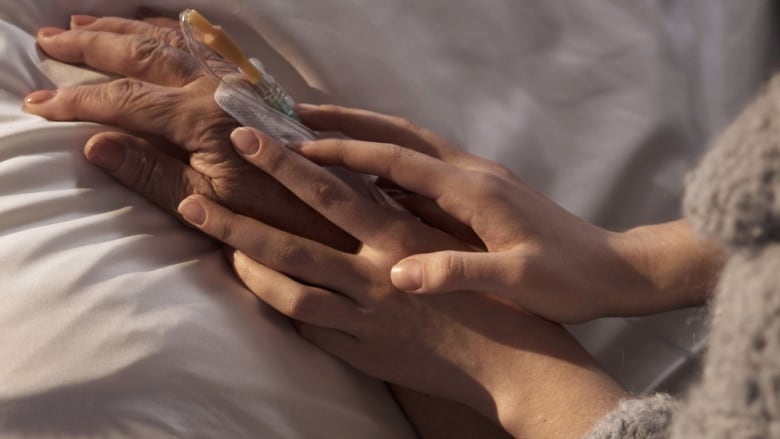 The hands of an adult hold the hand of an older person, who is hooked up to intravenous and lying in a bed.