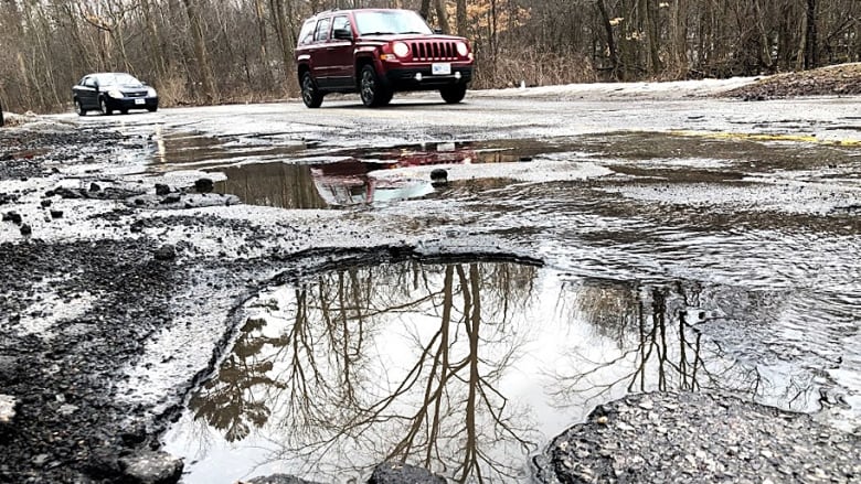 Cars drive by a large pothole filled with water.