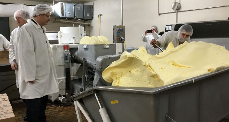 Workers clad in white jackets and hairnets shovel butter into a vat in a factory setting. 