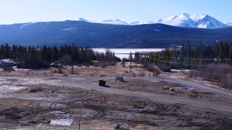 A view of an empty field with buildings, a lake and mountains in the background.