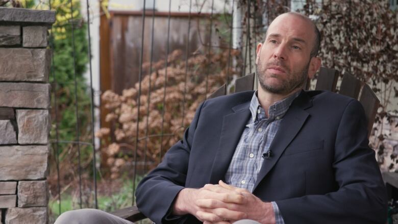 A white man in a suit and button-up shirt is sitting in an adirondack chair with his hands clasped on his lap. He has a thin beard and a buzz cut.