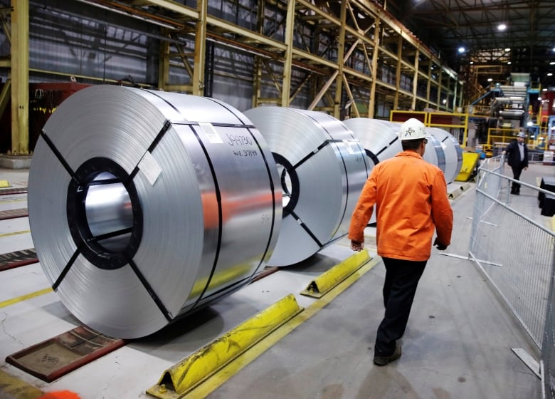 A man in safety equipment walks past rolled-up coils of steel.