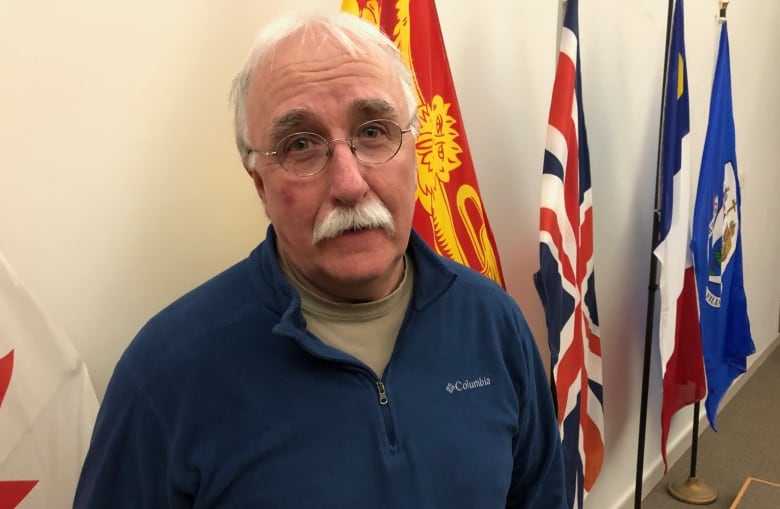 Man with short white hair and moustache and glasses wearing navy sweater stands in front of flags inside a building.