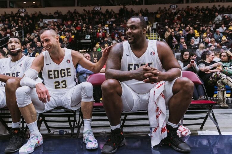 Two men smiling next to each other while sitting on a bench. The man on the left has his hand on the other man's shoulder.