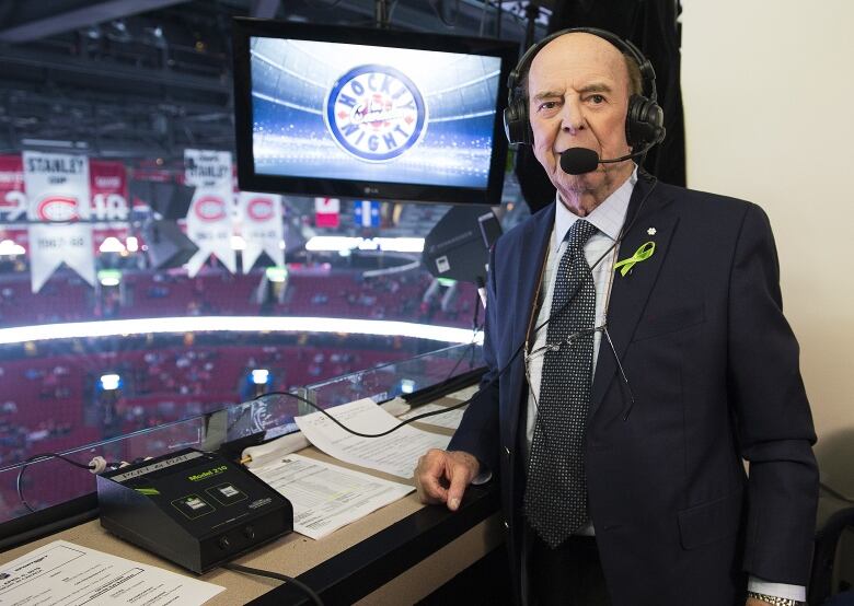 Older man wearing headset standing up in booth with hockey arena behind him.