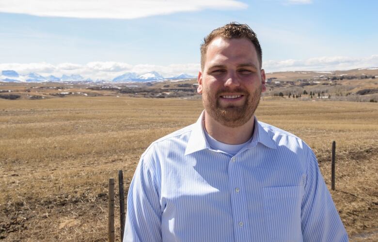 A man in a button up shirt stands in front of a field with mountains in the background. 