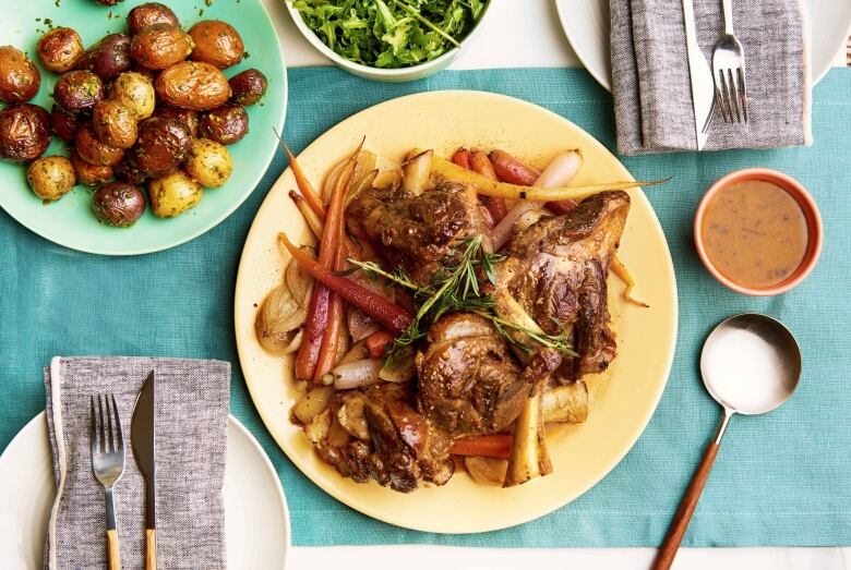 Overhead shot of braised lamb shanks sitting on a bed of roast vegetables on a plate. The plate is on a table that's set for a meal and a plate of roast potatoes sits next to the lamb shanks. 