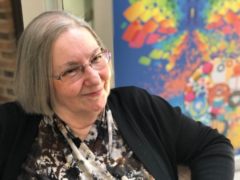 An older woman with grey hair and glasses sits in a chair and smiles.