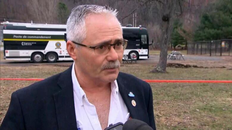 white man with grey hair in suit wearing pink collared shirt and navy jacket and square glasses standing near a crime scene outside.