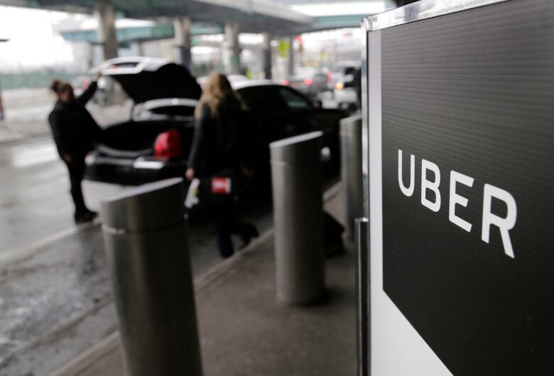 An Uber sign is in the foreground of the photo, in the background someone approaches a black vehicle while someone else loads it's trunk. 