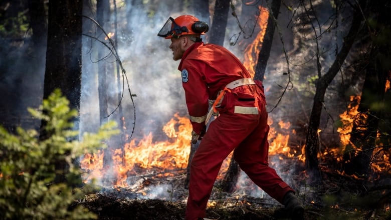 A firefighter in a red jumpsuit with soot on his face stands in smoke near a burning fire in a training exercise.