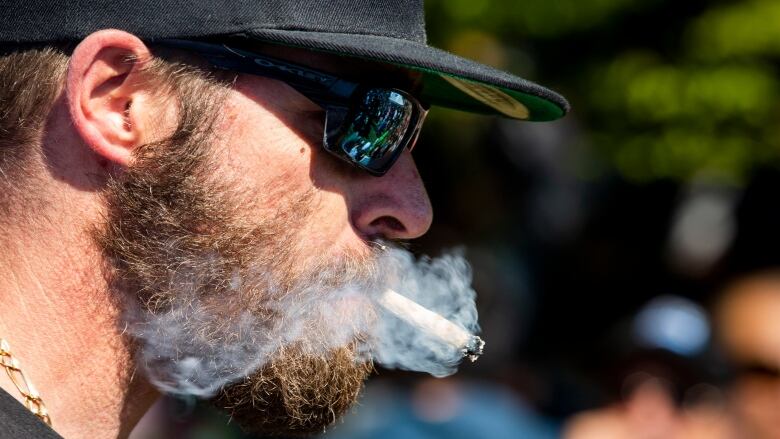 A man in a baseball cap and sunglasses shown in profile blows out a large cloud of smoke from the joint he is smoking.