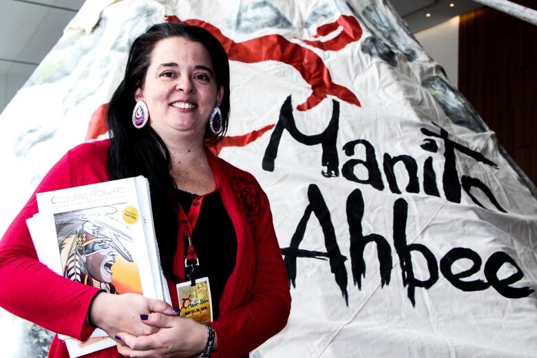 A woman with dark hair and red earrings wears a red sweater and smiles in front of a teepee