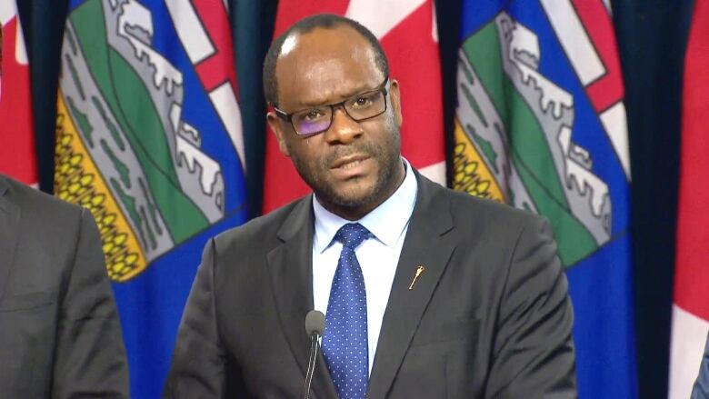 A man wearing glasses and a suit stands in front of Alberta and Canada flags.