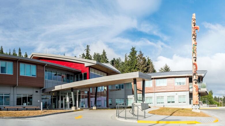 The front entrance of Haida Gwaii Hospital is a modern building with a large rectangular awning and a totem pole out in front of it.