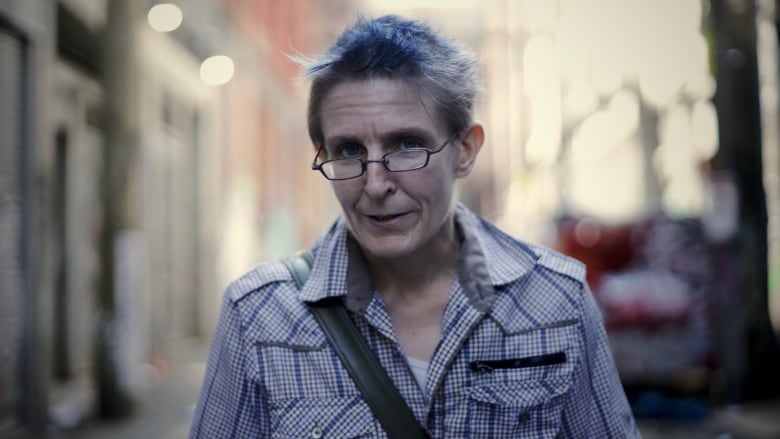 A woman wearing a checkered shirt and glasses stands on an urban street.